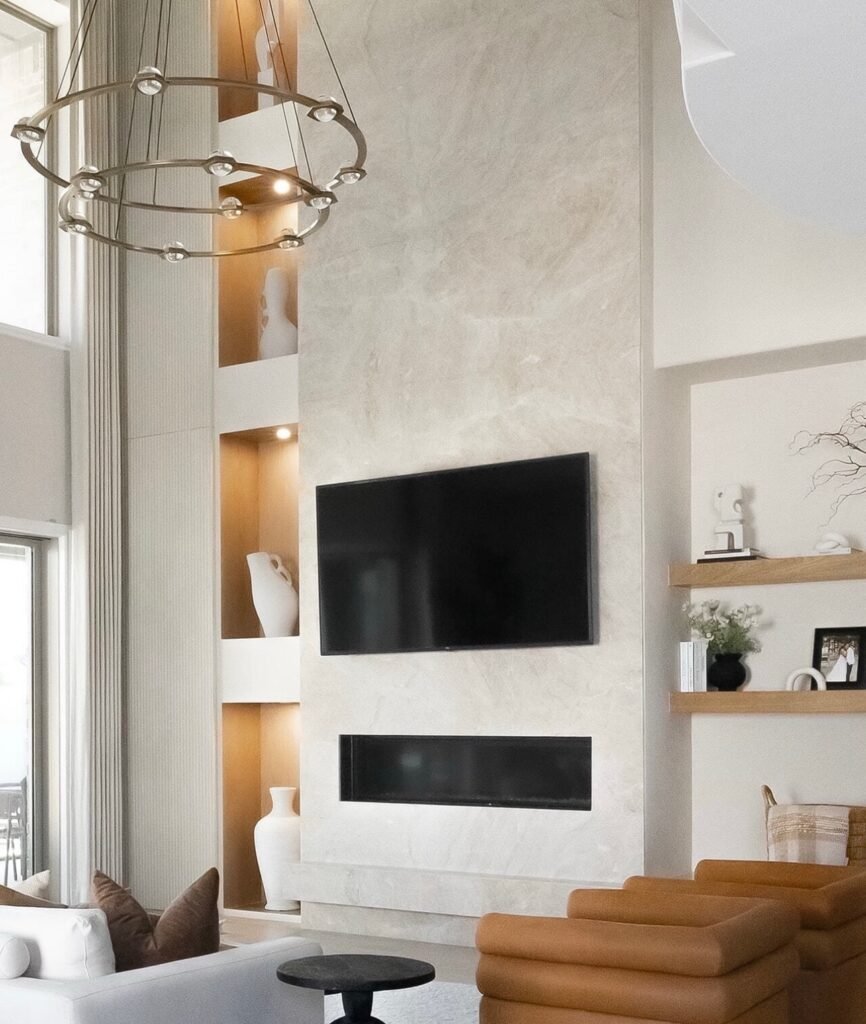 An open-concept living room featuring a floor-to-ceiling stone fireplace wall, mounted TV, and staircase view.